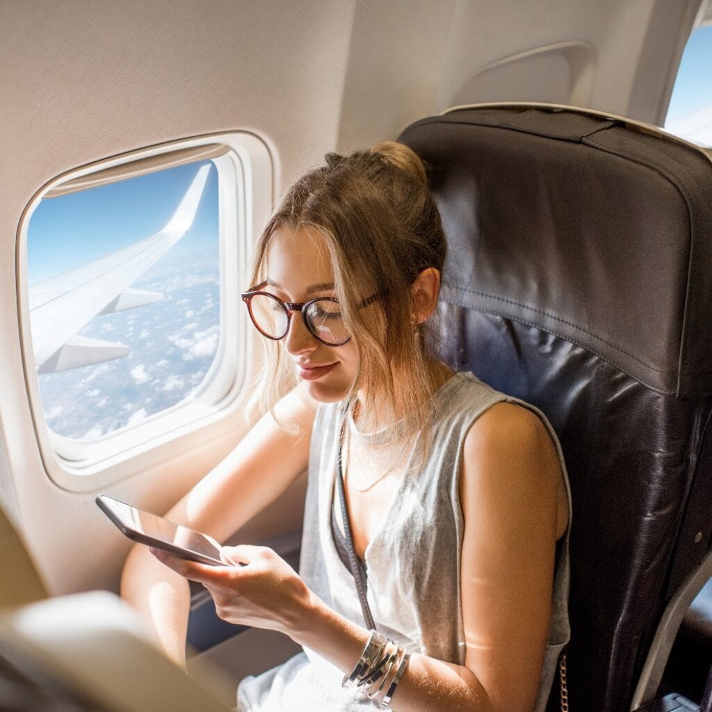 Young woman sitting with phone on the aircraft seat near the window during the flight in the airplane